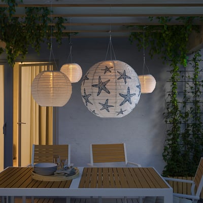 Four solar-powered pendant lamps, one with starfish design, illuminate outdoor dining area. They hang from pergola, casting warm glow.