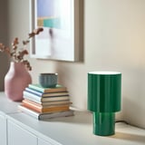 Small green SPETSBOJ lamp with ribbed design on table, near books, vase, and cup.
