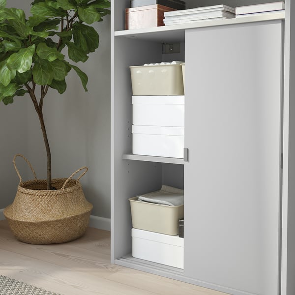 Open cabinet with stacked boxes and sliding door; plant and basket nearby.