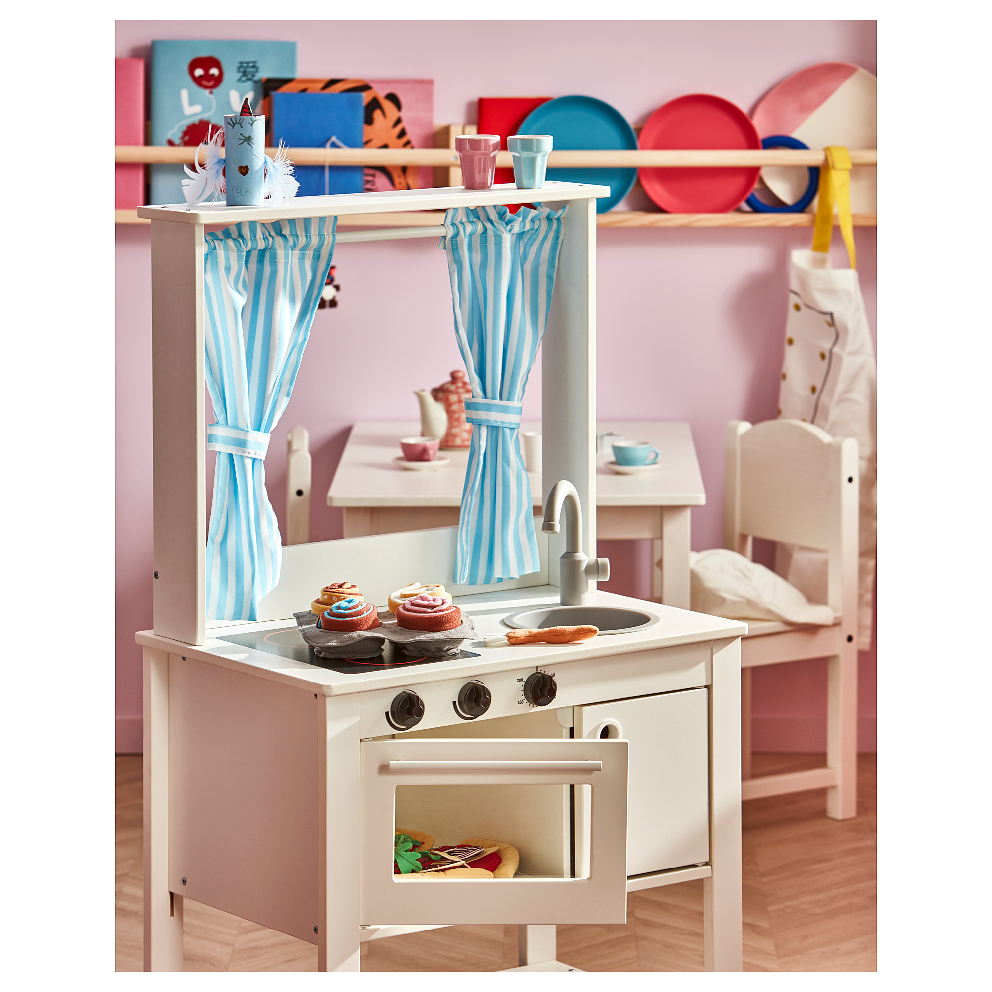 Childs white play kitchen with striped curtains, stove, sink, and toy food. Bright pink and blue accessories.