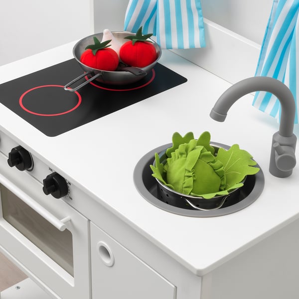 Toy kitchen with pretend food: tomatoes in pan, green leaves in sink. Black stove, gray faucet, white oven.
