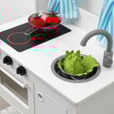 Toy kitchen with pretend food: tomatoes in pan, green leaves in sink. Black stove, gray faucet, white oven.