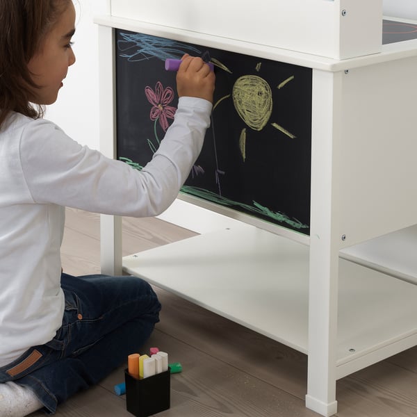 Child drawing on blackboard of white play kitchen, seated on floor using colored chalk.