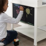 Child drawing on blackboard of white play kitchen, seated on floor using colored chalk.