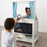 Children playing with play kitchen, one serving, blackboard menu, curtains.