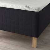 Close-up of a modern bed base with wooden legs and a white mattress by ikea. The base has a padded dark cover with a grid pattern.