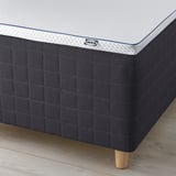 A corner of an IKEA mattress with a gray quilted base and wooden legs on a wooden floor.