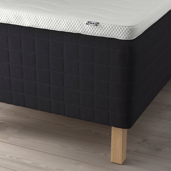 Image shows a corner of a black IKEA mattress on wooden legs. The mattress has a quilted pattern and a white top layer with the IKEA logo visible.