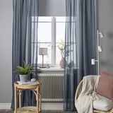 Dark blue curtains frame large window. Plant, book on table; chair with blanket, pillow and nearby light.