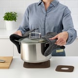 A person uses a STENPOTT trivet to lift a pots lid on a countertop.