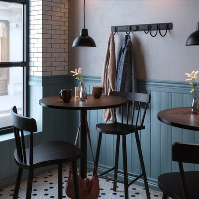Modern café setting with round dark tables, high black chairs, and hanging mugs. Blue-green panel walls and brick accents.