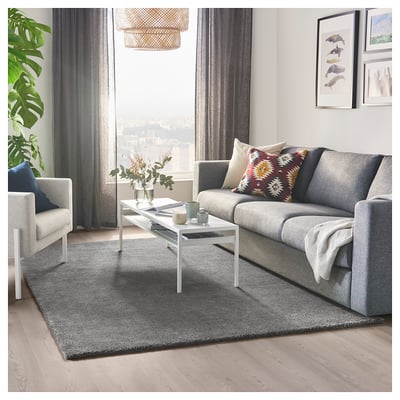 Modern living room with grey STOENSE rug, grey sofa, white coffee table, colorful throw pillows, and large windows with city view.