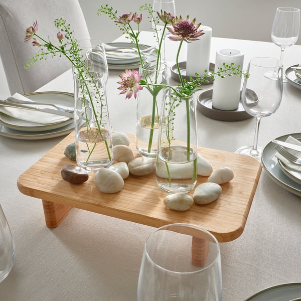Wooden raised platform displaying three glass vases with purple flowers and pebbles, surrounded by plates, glasses, and candles on a table.