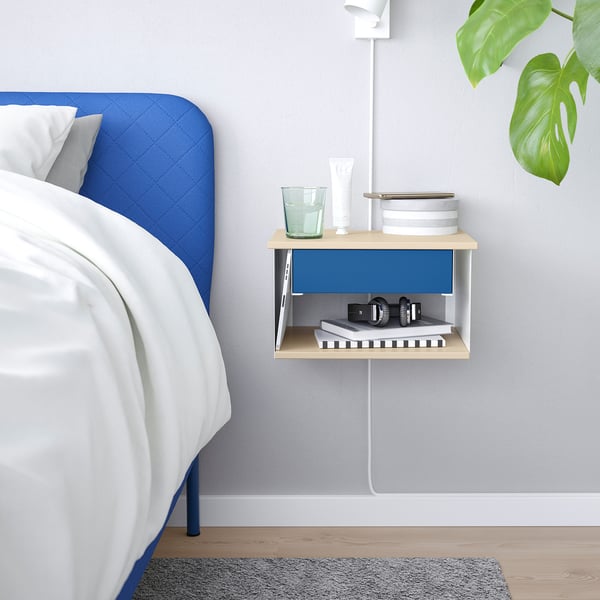 Wall-mounted STOMSÖ nightstand in blue and wood with open/closed storage, holding glass, books, lamp, and plant.