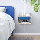 Wall-mounted STOMSÖ nightstand in blue and wood with open/closed storage, holding glass, books, lamp, and plant.
