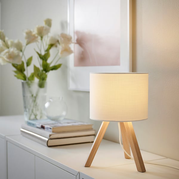 Small white STORSEGEL lamp with wooden tripod base, next to flowers and books, warm light.