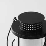 Black lantern with metal handle, featuring a perforated design for ventilation and a glass cover at the bottom, standing against a white background.