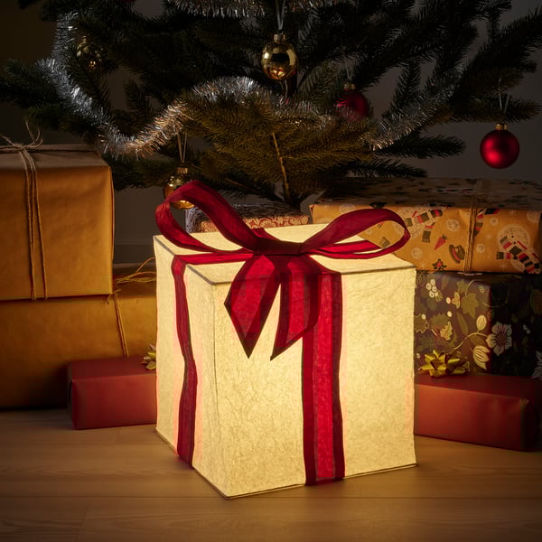 Small glowing cube light, cream-colored with red ribbon, under a christmas tree, surrounded by wrapped gifts.