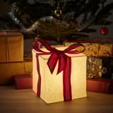 Small glowing cube light, cream-colored with red ribbon, under a christmas tree, surrounded by wrapped gifts.