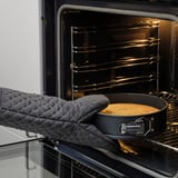 A baker uses oven mitts to place a golden-brown baked good into an oven, showcasing STRANDFLY pans even heat distribution and non-stick properties.