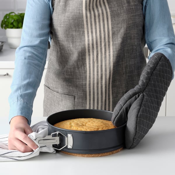 Person holding a round, baked dish with a springform pan, using oven mitts. The non-stick pan has a lock mechanism for easy removal of baked goods.