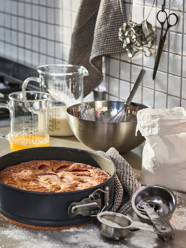 Black springform pan with golden-brown baked dessert, surrounded by glass measuring cups, metal bowl, utensils, and bags, on a kitchen counter.