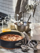 Black springform pan with golden-brown baked dessert, surrounded by glass measuring cups, metal bowl, utensils, and bags, on a kitchen counter.