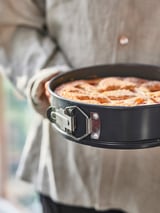 A black springform pan with a golden-brown baked dish, held by a person in an apron.
