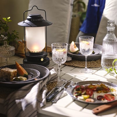 Table set with modern floral porcelain, STRIMMIG. Centered lantern provides warm light. Plates hold fresh food. Glasses and decanter suggest drinks.