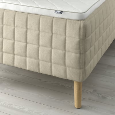 Close-up of a STUVLAND mattress edge with natural linen texture and wooden legs on light wood floor.