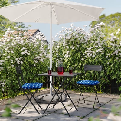 Small patio set with folding table and blue-cushioned chairs, shade by white umbrella, set against a wall of blooming white flowers.