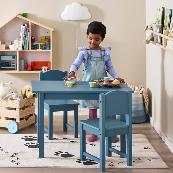 Kid in overalls playing with toy cups on blue kids table. Room has paw rug and padded toy box.