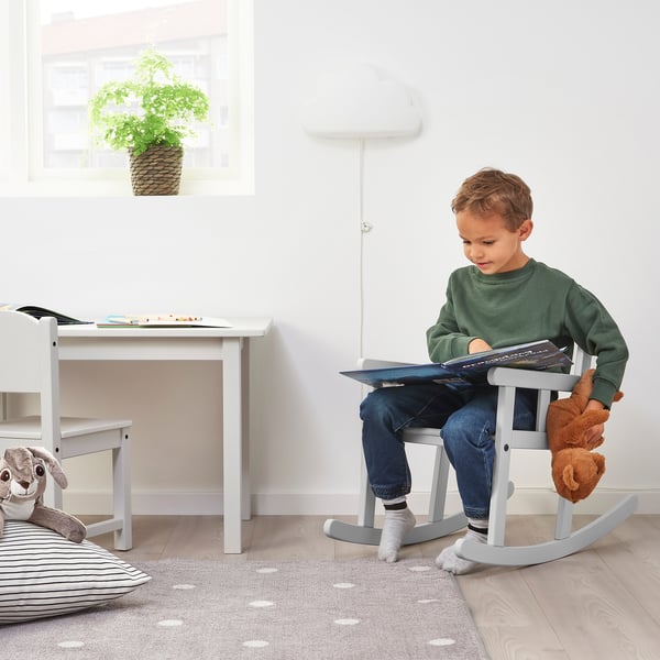 Boy reads at childs desk with stuffed animal. Nearby table has books and puzzles. Striped cushion and toy on floor, plant on window.