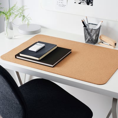 Beige cork desk pad on white desk, with notebooks, fern, pen holder, and stapler.