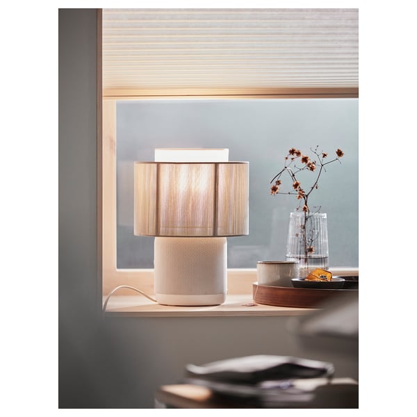 Modern minimalist lamp with wifi speaker. White, cylinder shape with textured surface and beige lamp shade. Placed on window sill with decor.