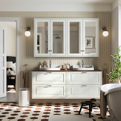 Modern bathroom with double-sink vanity in white. Large mirror, wall lamps, patterned tile floor. Plants and towels add decor.