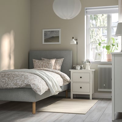 Cozy bedroom with gray TÄRNKULLEN bed, padded headboard, and wooden legs. Neutral bedding, nightstand with lamp, and large window with plant.