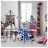 Two women collaborate in a colorful studio. One sews while the other assists, surrounded by vibrant fabrics.