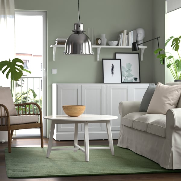 Modern living room with green walls, green rug, white table, gray chair, wicker chair, and decorator shelves.