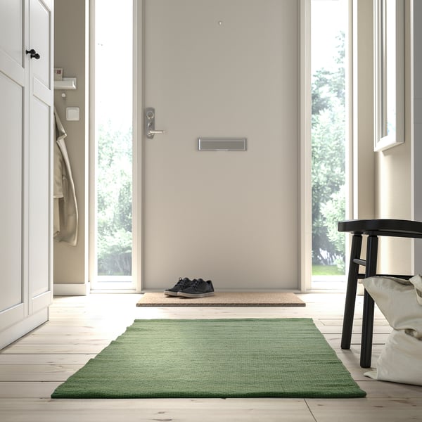 A green TIDTABELL rug, rectangular, flatwoven with wool surface, placed in a hallway beside a black stool and near a door.