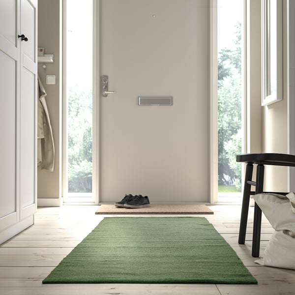 A TIDTABELL green rug placed near a door, smaller than the doorway and a nearby chair.