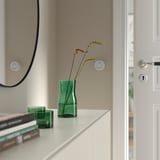 Green glass vase with flowers on shelf, books, mirror, door.