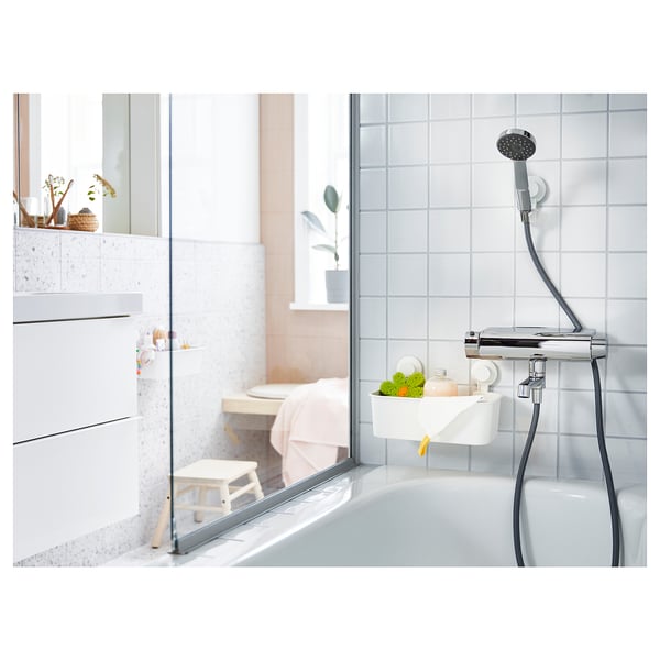 Modern white bathroom with glass shower, chrome fixtures, and a wooden step stool. TISKEN wall-mounted basket holds fruits next to shower head.