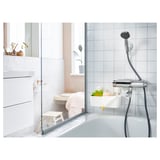 Modern white bathroom with glass shower, chrome fixtures, and a wooden step stool. TISKEN wall-mounted basket holds fruits next to shower head.