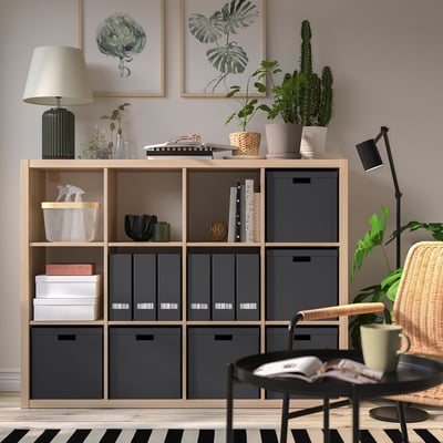 Modern living space with TJENA storage, dark grey boxes, books, plants, wicker chair, black coffee table.