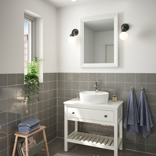 Modern bathroom with TOFTBYN white mirror above white vanity. Gray tiled walls, hanging blue towels, and potted plant.