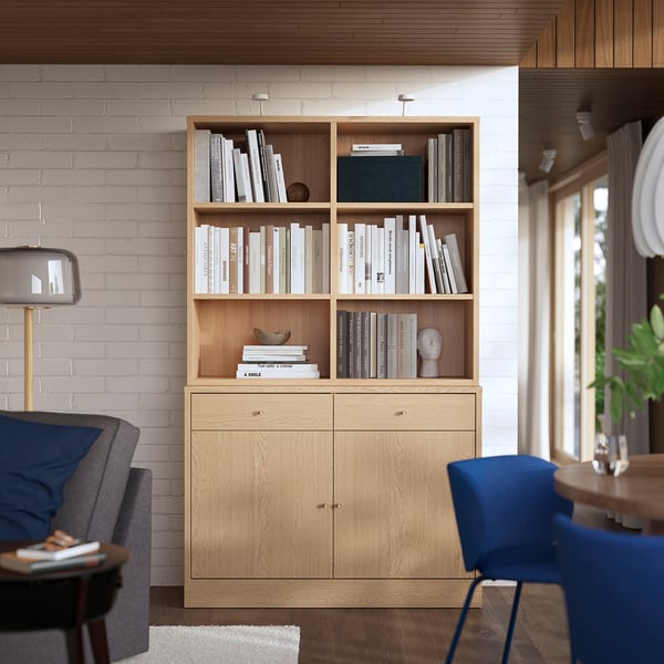 Wooden bookshelf with open & closed storage, filled with books. Against white brick wall, next to grey chair & blue stool.