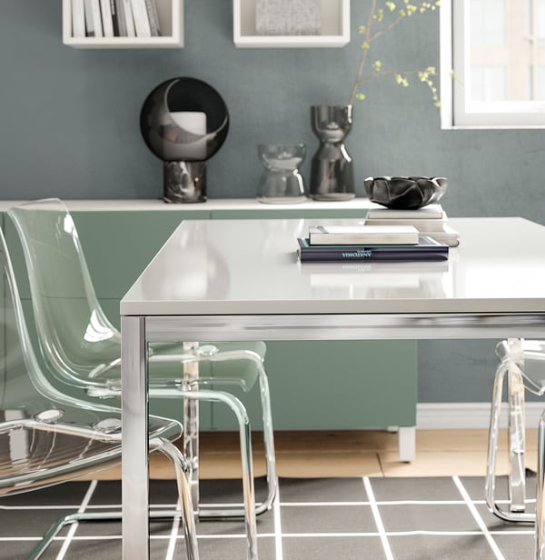 Modern glass table with metallic legs, green chairs, decorative items on a shelf, and a blue wall.
