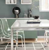 Modern glass table with metallic legs, green chairs, decorative items on a shelf, and a blue wall.