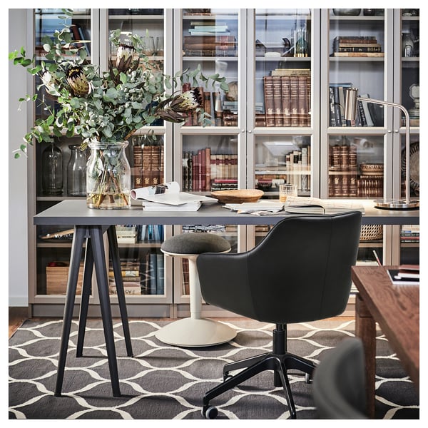 Modern home office setup with TOSSBERG black leather swivel chair, gray desk, floral vase, patterned rug, and glass cabinet filled with books.